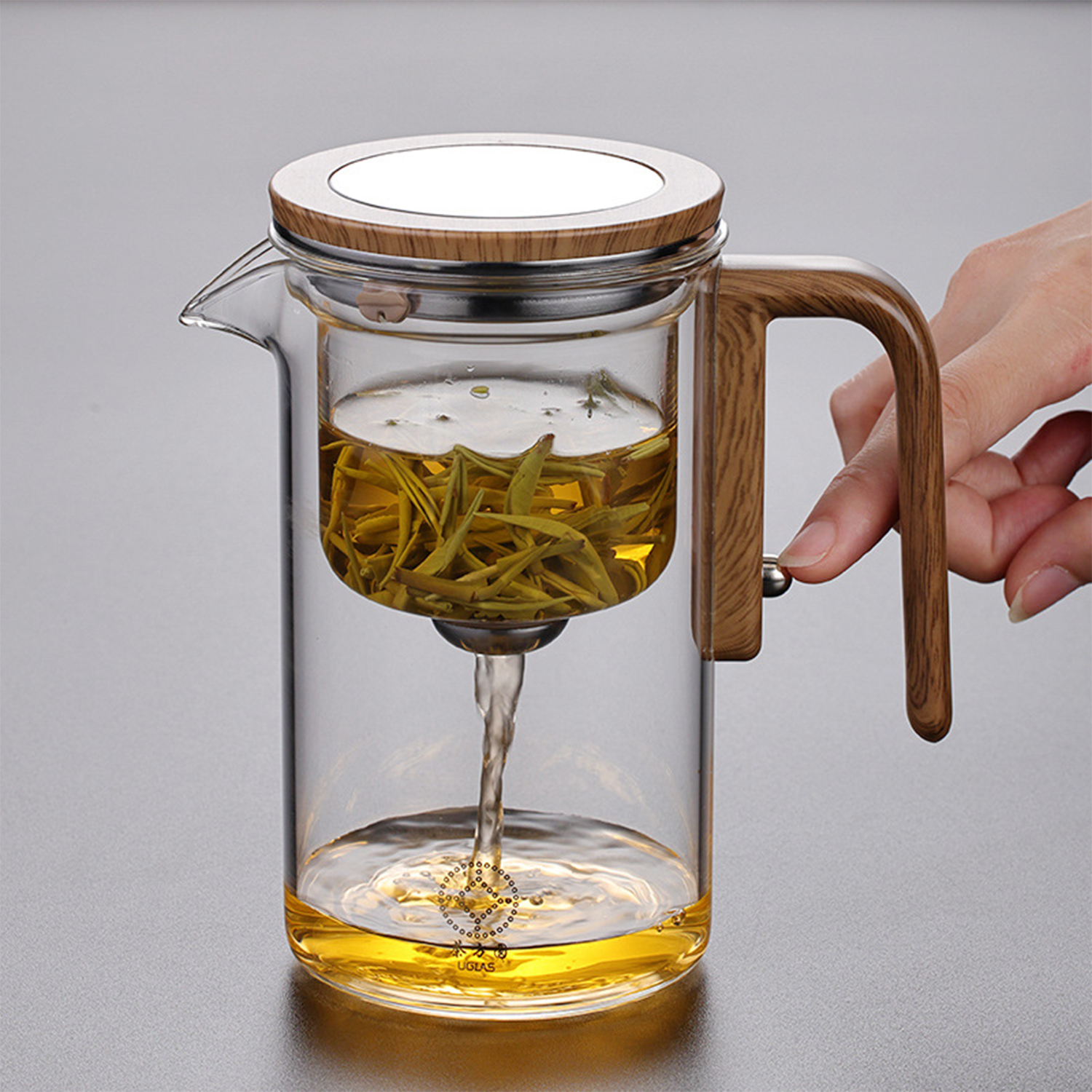 Una mano presiona el botón de una tetera de vidrio con detalles de madera mientras el té fluye hacia abajo.