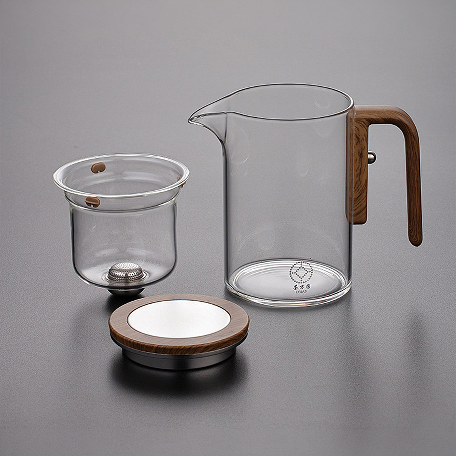 Una tetera de vidrio transparente con mango de madera, un colador a juego y una tapa redonda de madera sobre fondo gris.