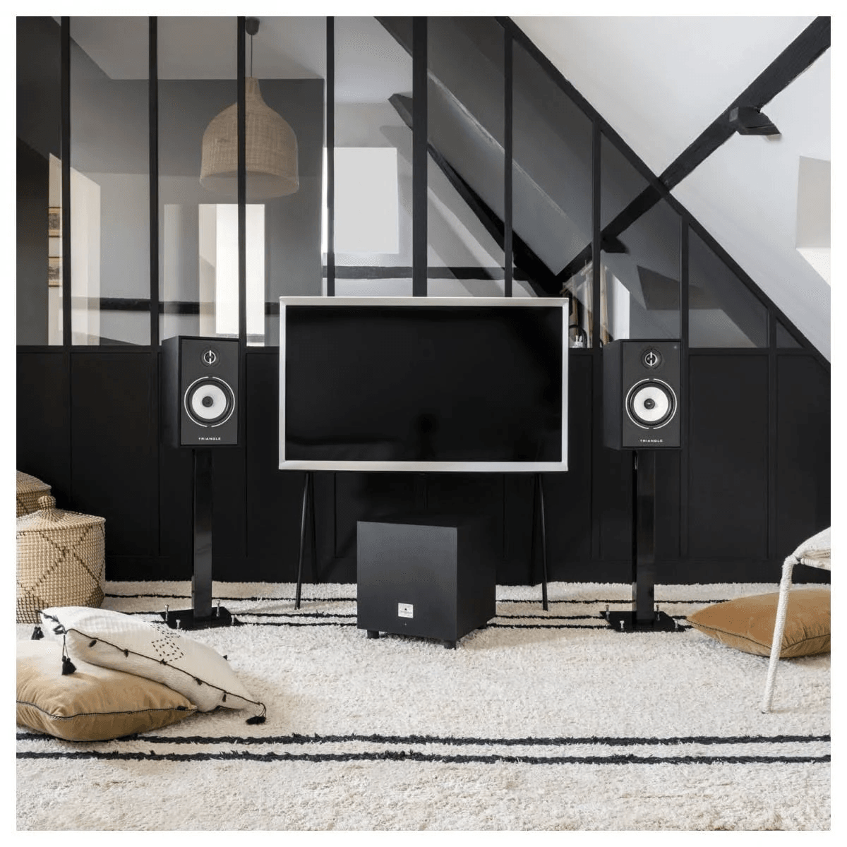 Una sala con televisor, dos altavoces, un subwoofer, alfombra y cojines frente a una pared de vidrio y negro.