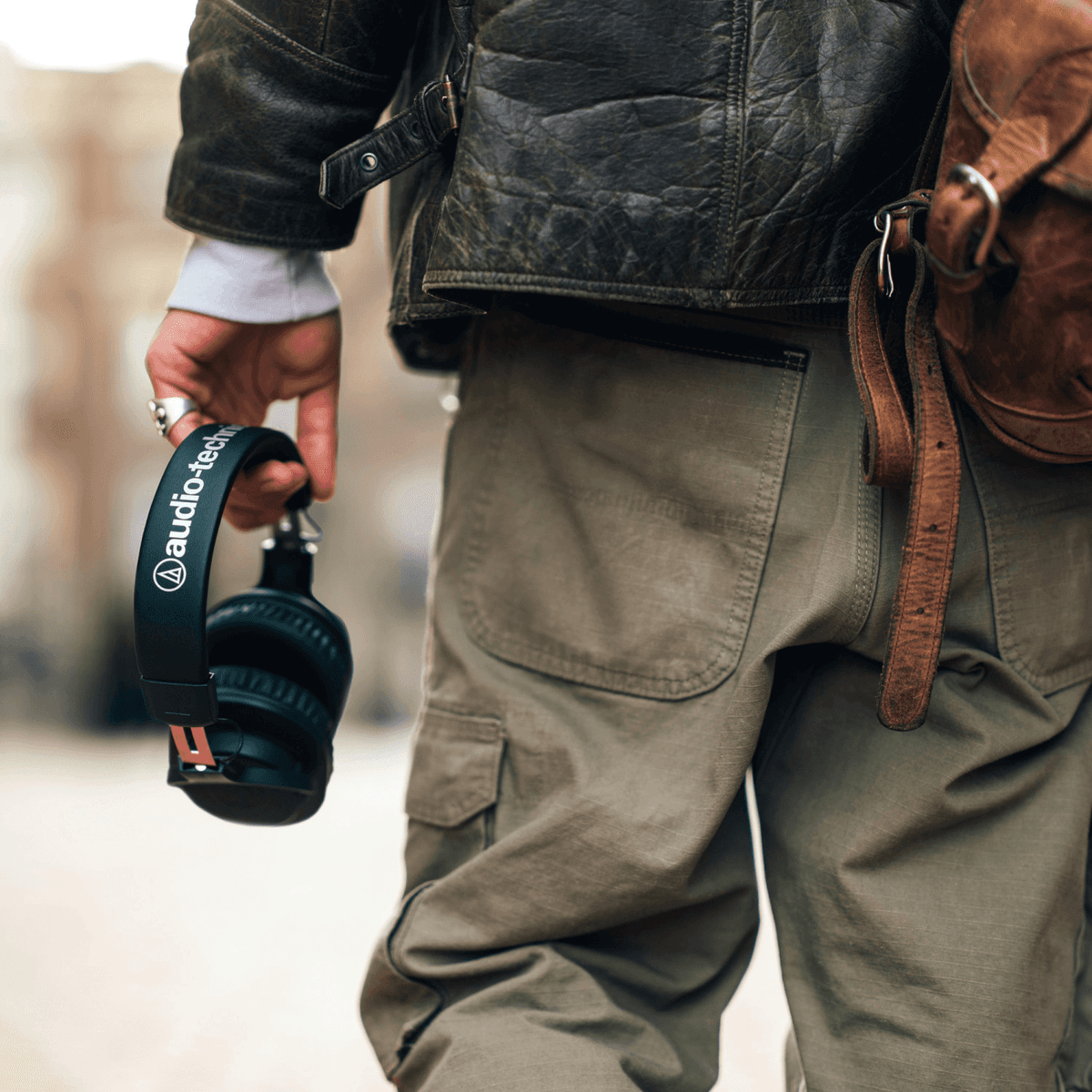 Una persona con giacca di pelle e pantaloni cargo tiene cuffie nere Audio-Technica, con borsa di pelle.