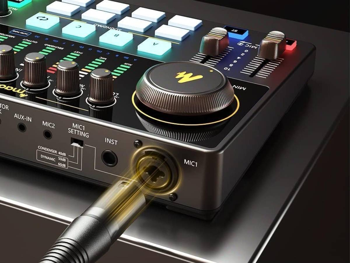 Primo piano di una console di missaggio audio nera con manopole, fader e un cavo per microfono collegato a MIC1.