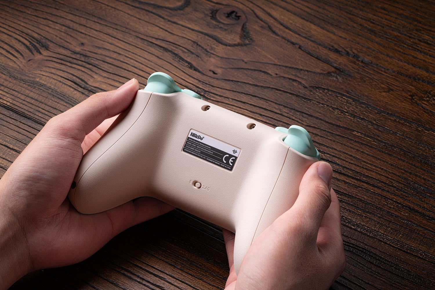 Mani che tengono un controller wireless 8BitDo beige con pulsanti verde menta su un tavolo di legno.