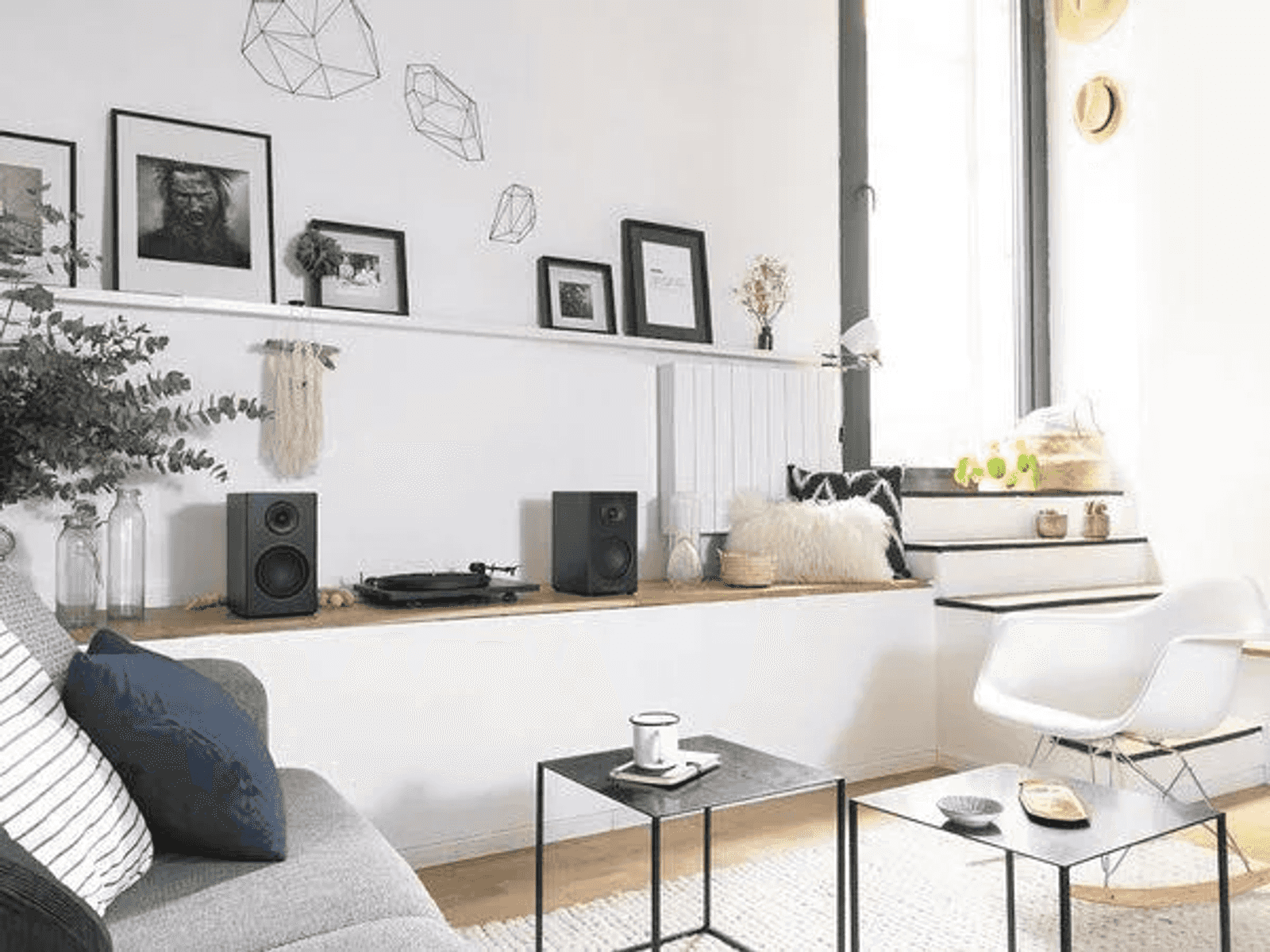 Una sala de estar luminosa con pared blanca, altavoces negros, cuadros enmarcados y dos mesas negras pequeñas.