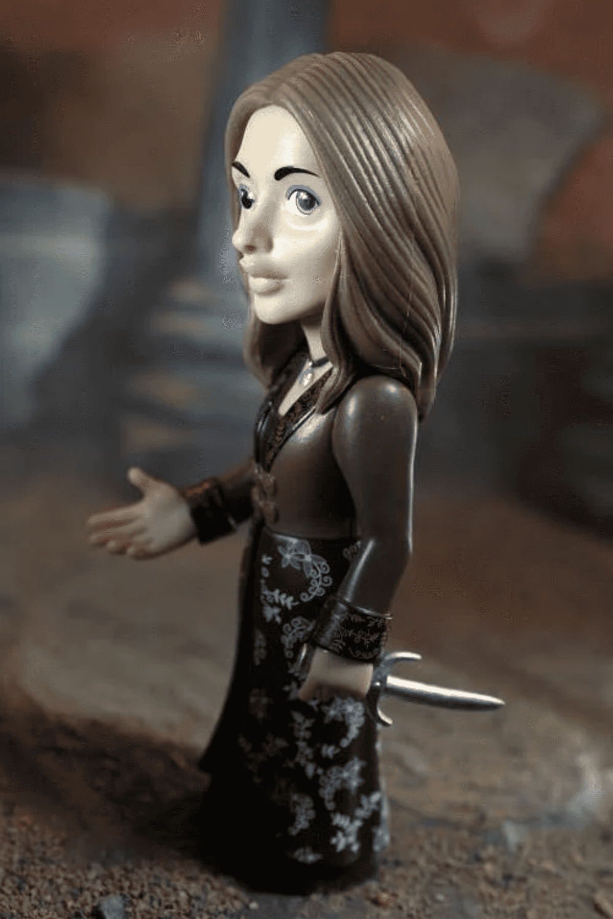 Un'action figure femminile con i capelli castani indossa un abito scuro floreale e tiene un pugnale.