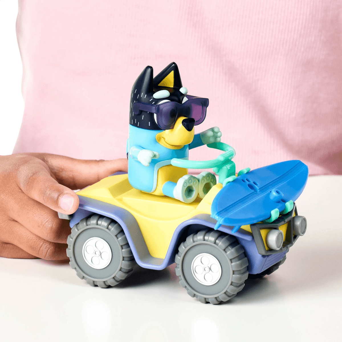Bluey-Spielfigur mit Sonnenbrille auf einem bunten Spielzeug-Quad mit Surfbrett.