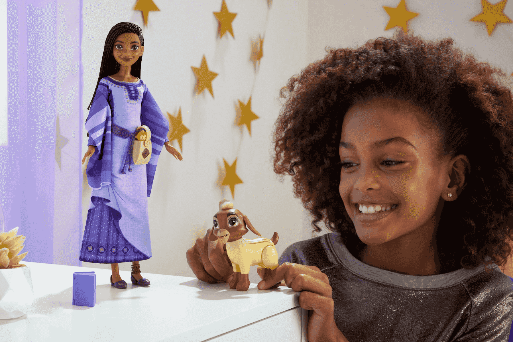 Ein smiling Mädchen mit Afro-Haaren spielt mit einer Puppe und einer Ziegenfigur vor einer Wand mit Sternen.