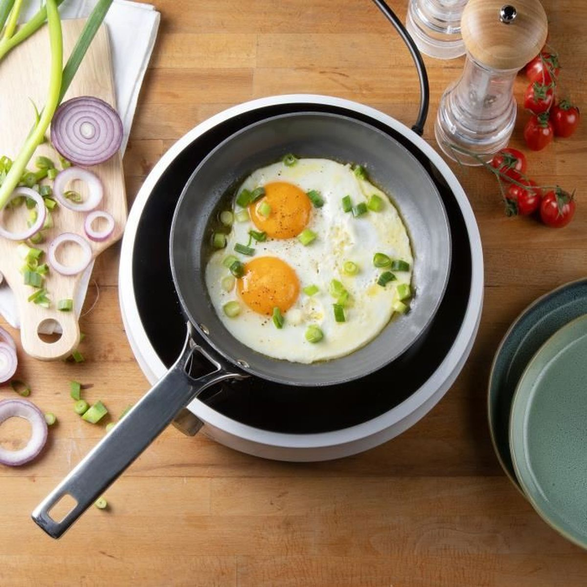 Zwei Spiegeleier mit Frühlingszwiebeln in einer Pfanne auf einer Herdplatte mit Gemüse daneben.