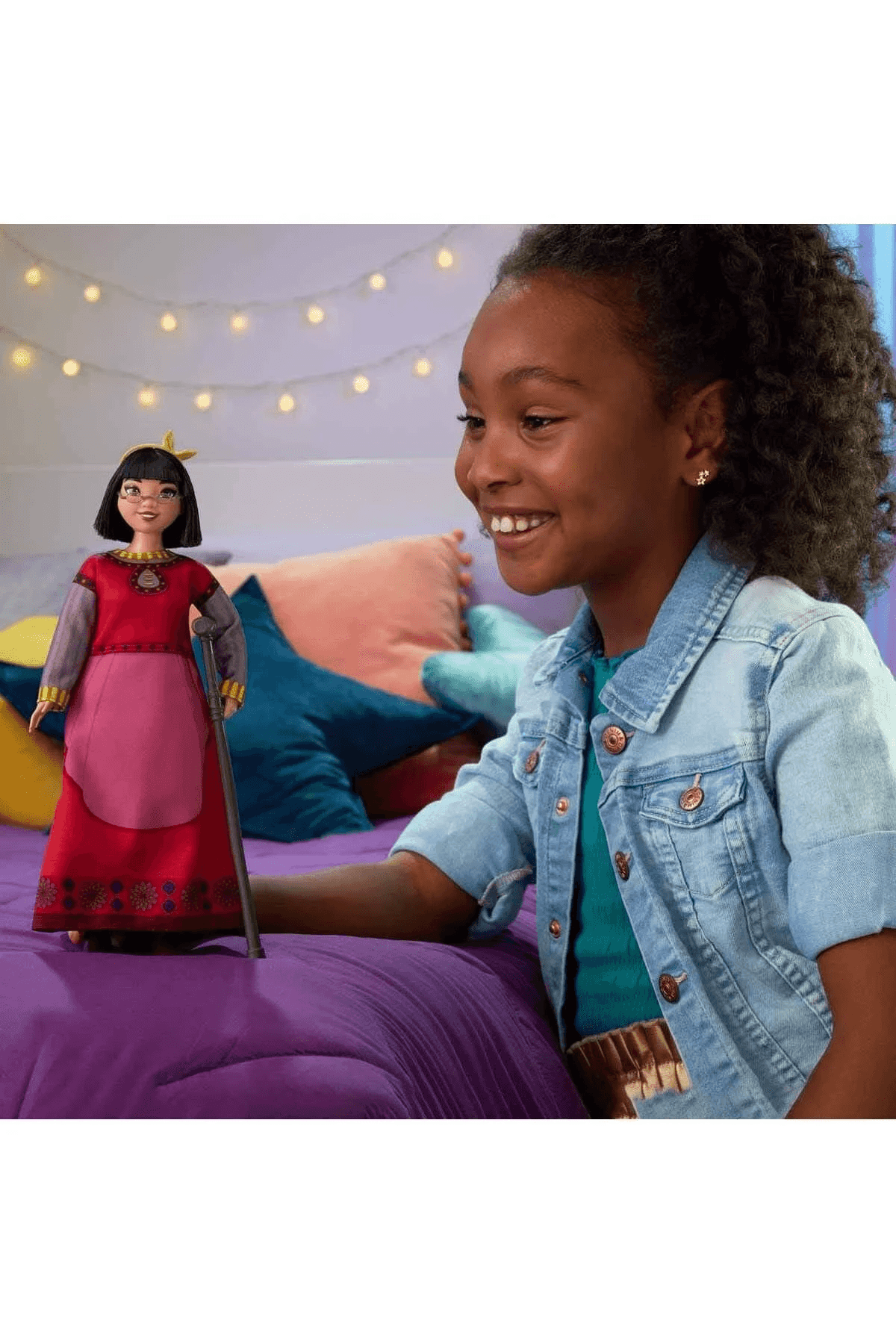 Ein smiling Mädchen mit Denim-Jacke hält eine Puppe mit rotem Kleid auf einem lila Bett.