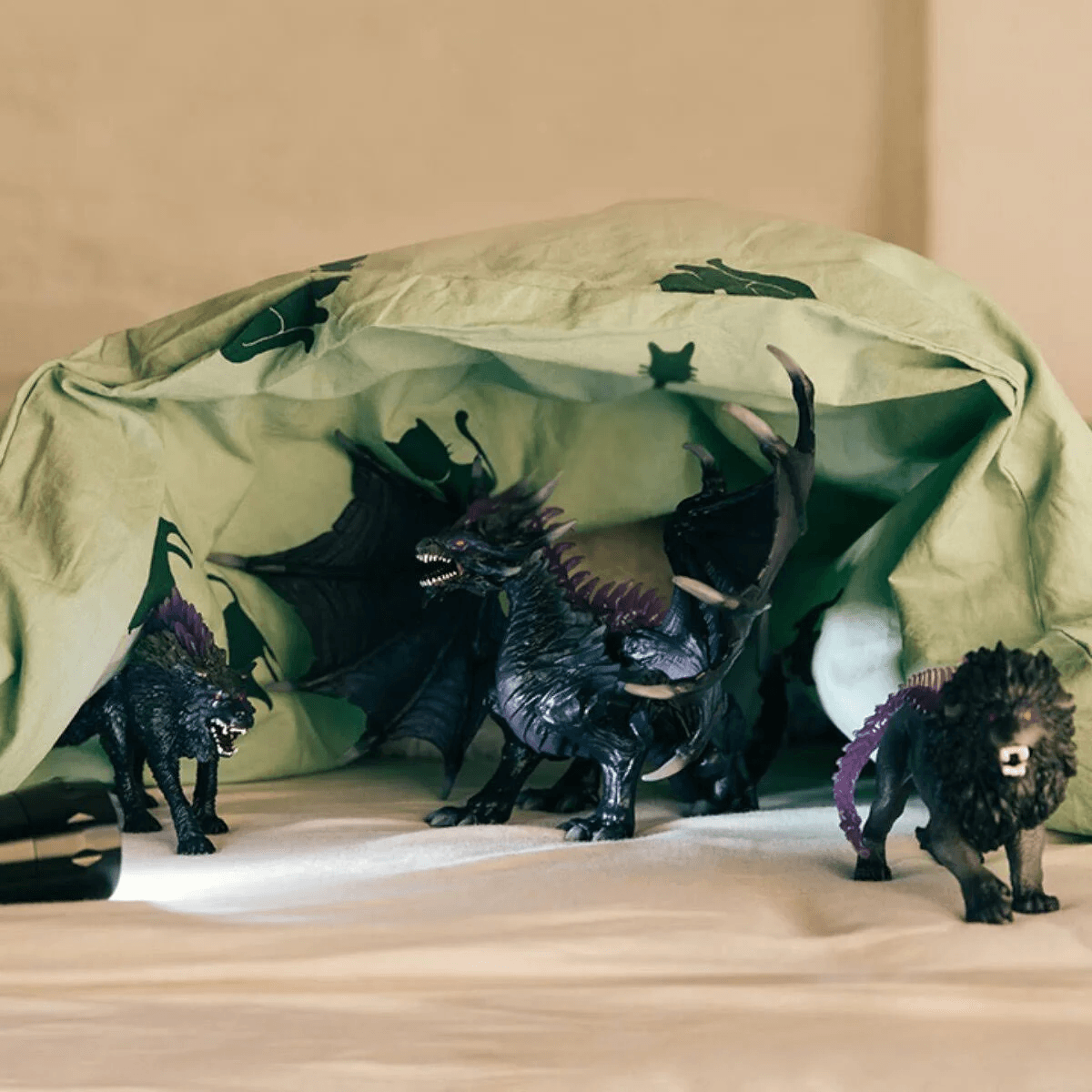 Schwarze Spielfiguren von einem Drachen, einem Wolf und einem Löwen gucken unter einer grünen Decke hervor.