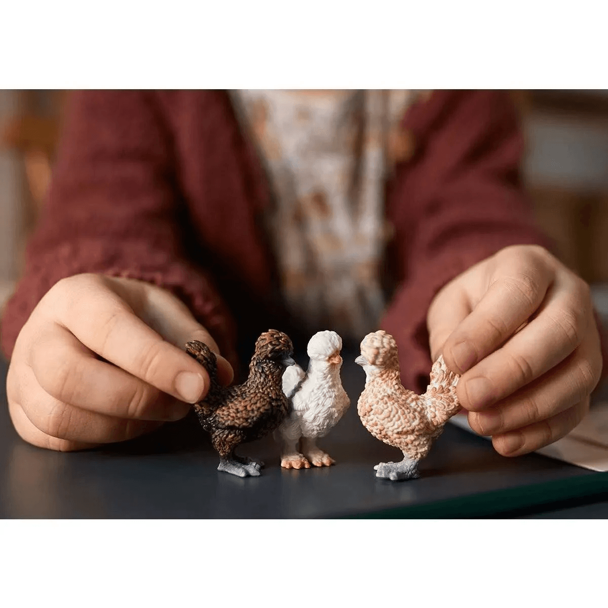 Ein Kind hält drei kleine, strukturierte Hühnerfiguren in braun, weiß und beige auf einem Tisch.