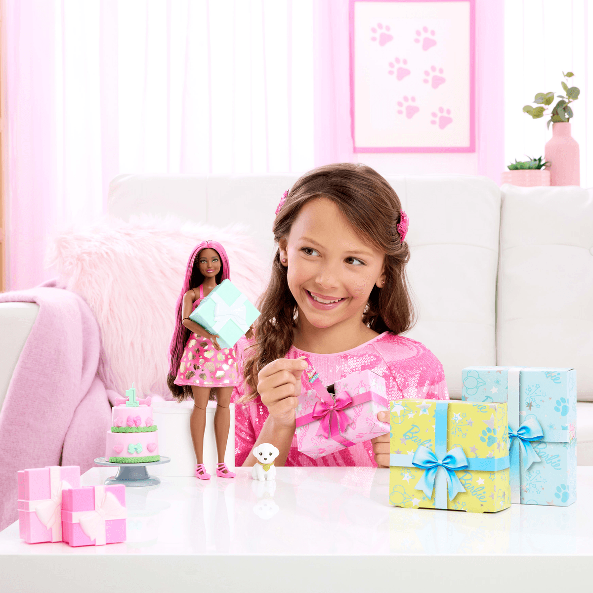 Una bambina sorridente apre un regalo rosa su un tavolo con pacchetti, una bambola Barbie e un cane giocattolo.