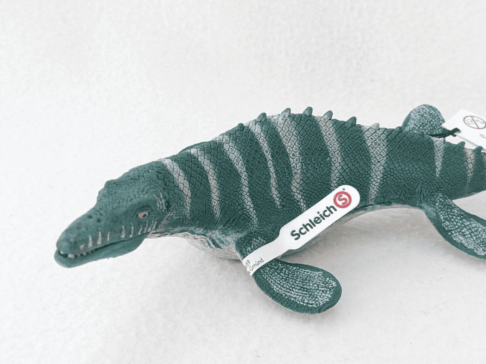 Un giocattolo Mosasauro Schleich con pelle squamosa verde e etichetta su superficie bianca.