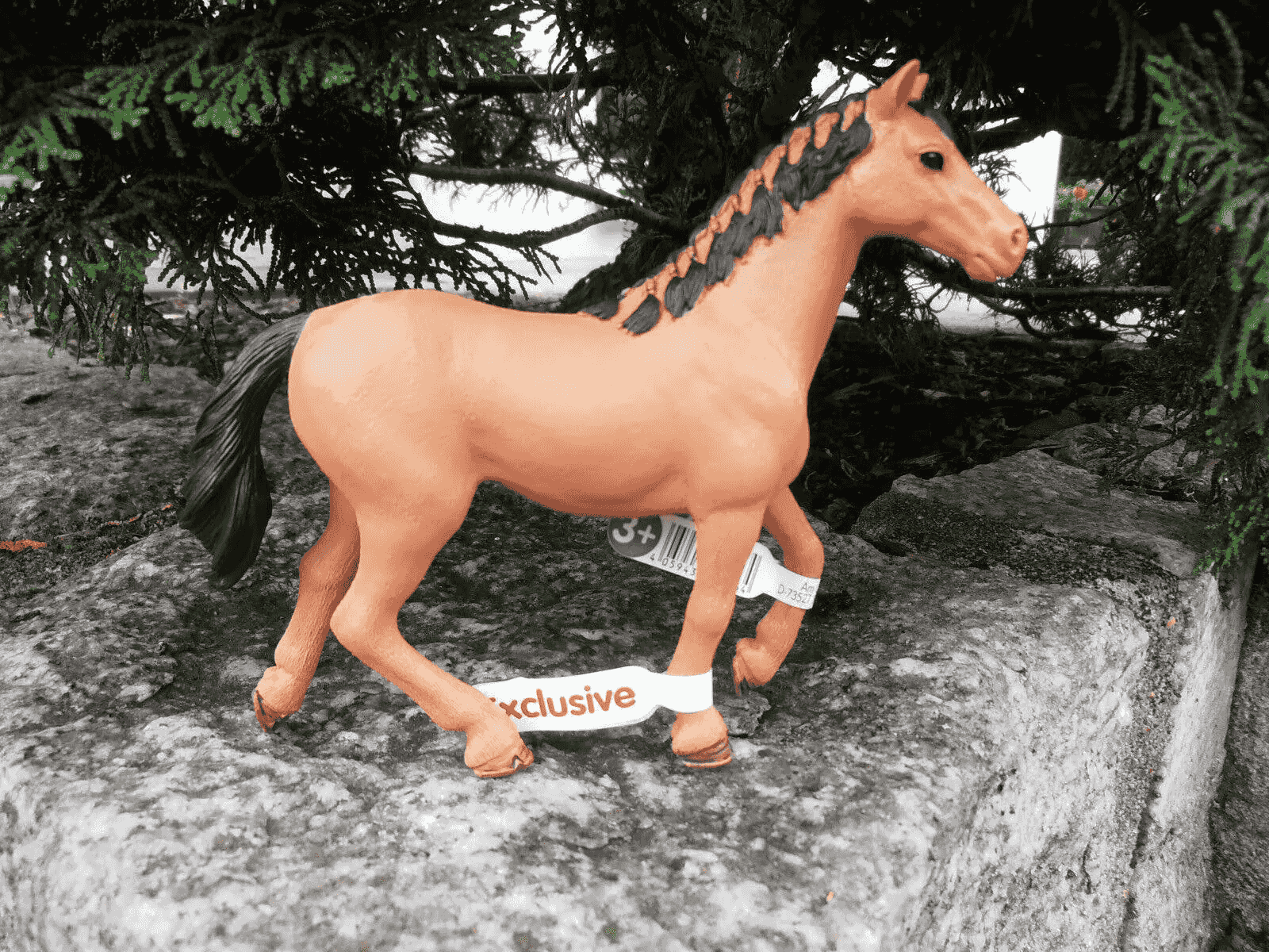 Una figura de caballo de juguete marrón con crin y cola negras sobre una piedra bajo ramas de pino.