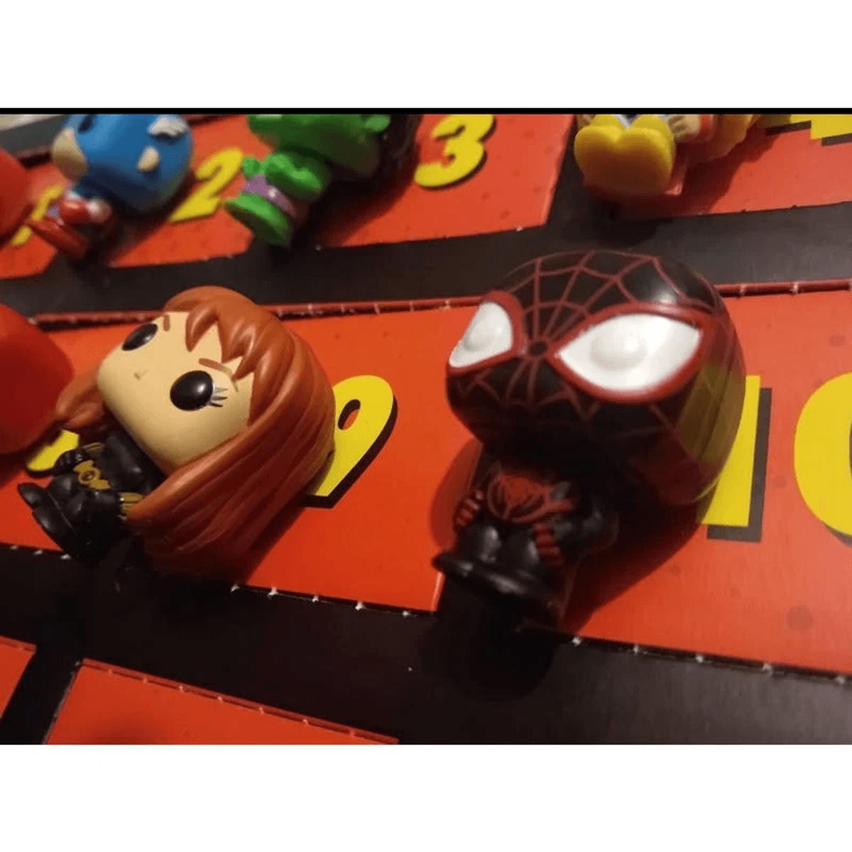 Diverse statuine in vinile di supereroi, tra cui Spider-Man e Black Widow, giacciono su una superficie rossa numerata.
