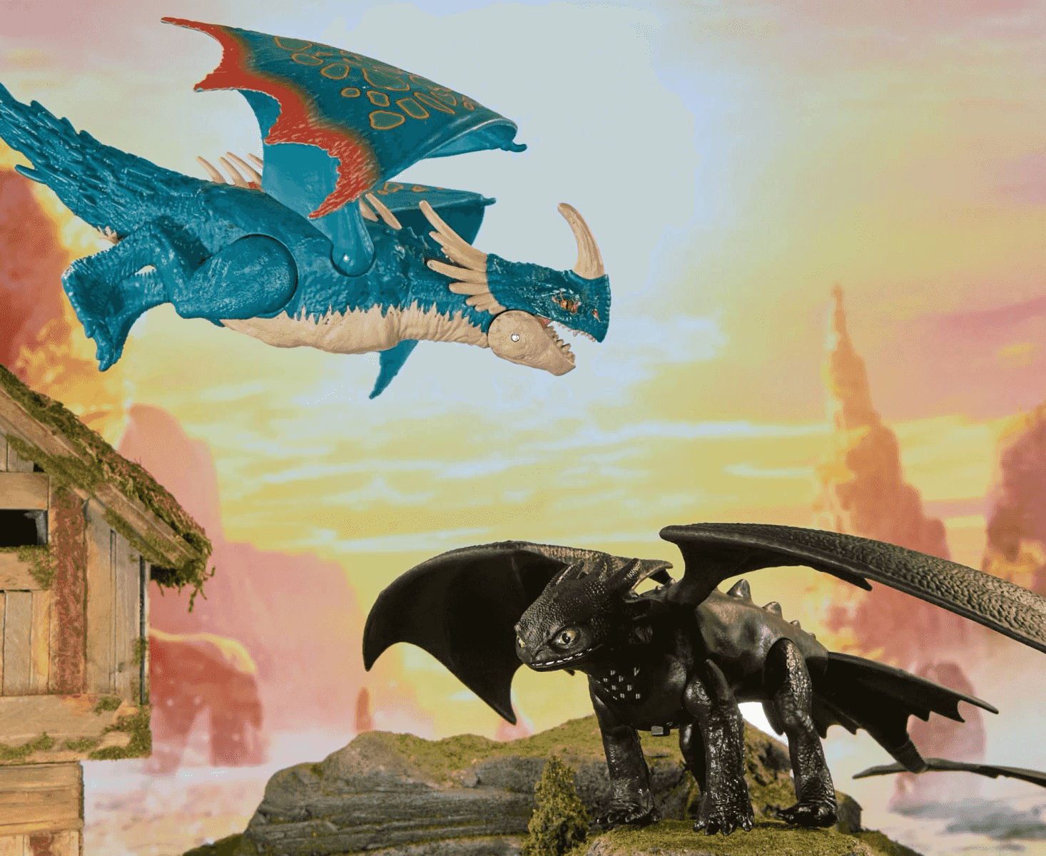 Dos juguetes de dragón, uno azul volando y otro negro de pie en una colina rocosa con fondo de cielo al atardecer.