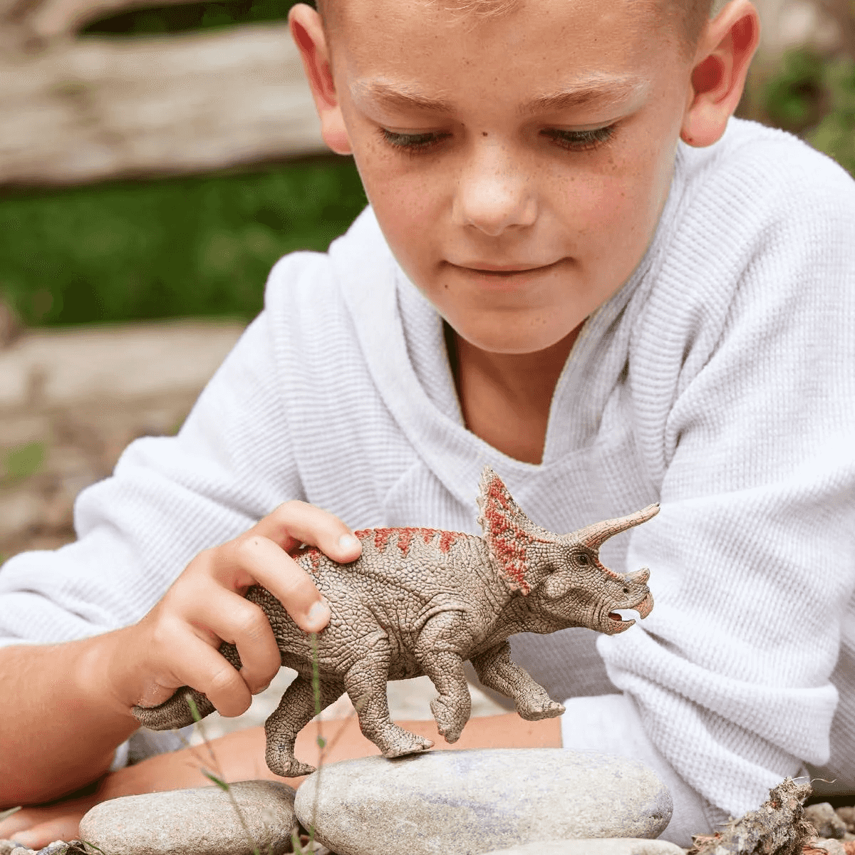 Un ragazzo con un maglione bianco gioca con un dinosauro triceratopo giocattolo sulle rocce.