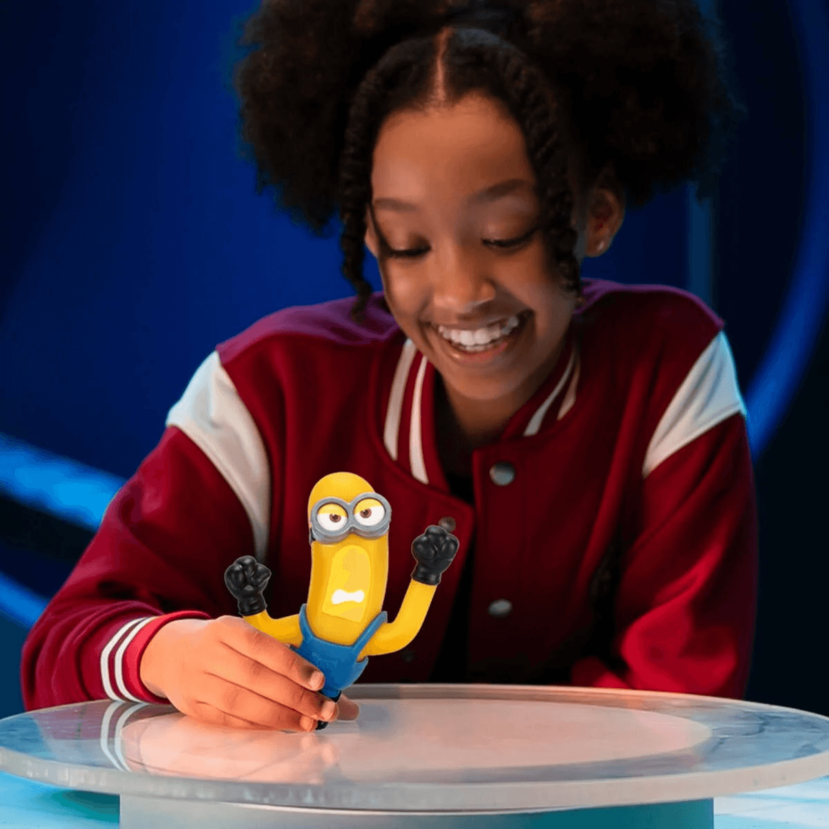Una niña sonríe mientras sostiene una figura de juguete de Minion en una mesa de vidrio.