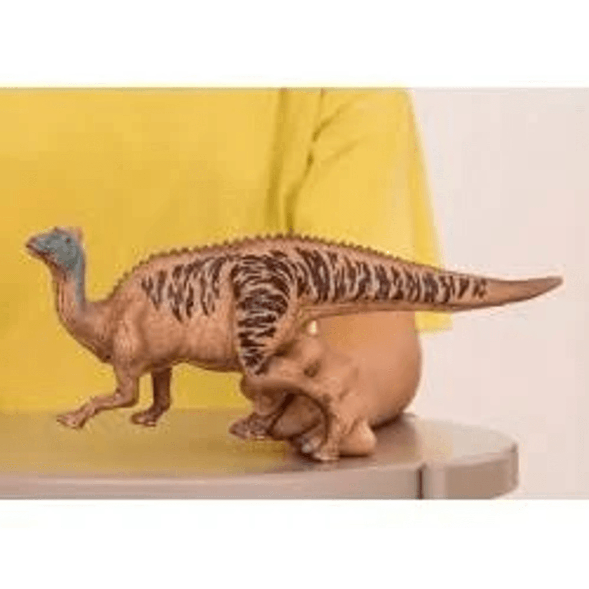 Una mano tiene un giocattolo di dinosauro marrone con strisce scure su un tavolo davanti a uno sfondo giallo.