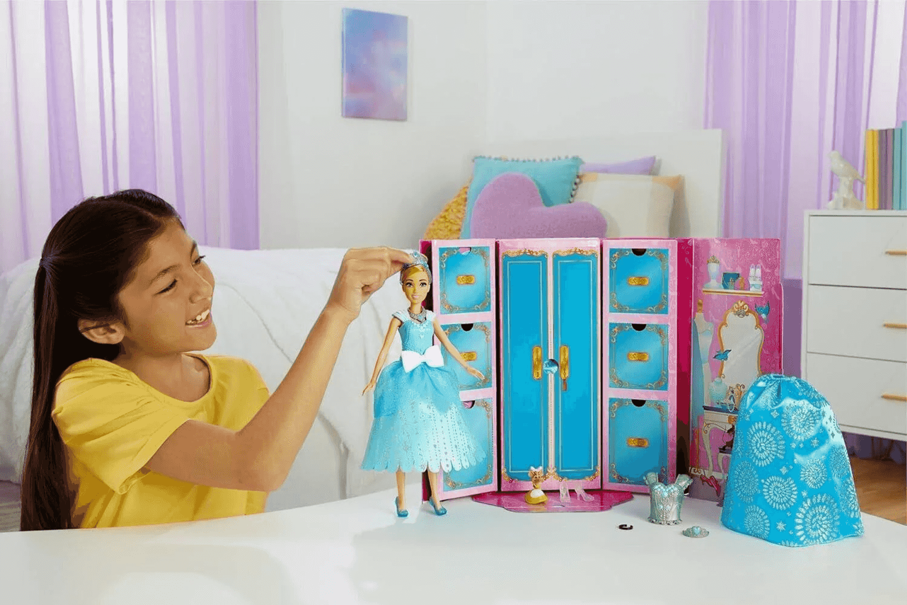 Ein junges Mädchen im gelben Shirt spielt vor einem blau-rosa Kleiderschrank mit einer Cinderella-Puppe.