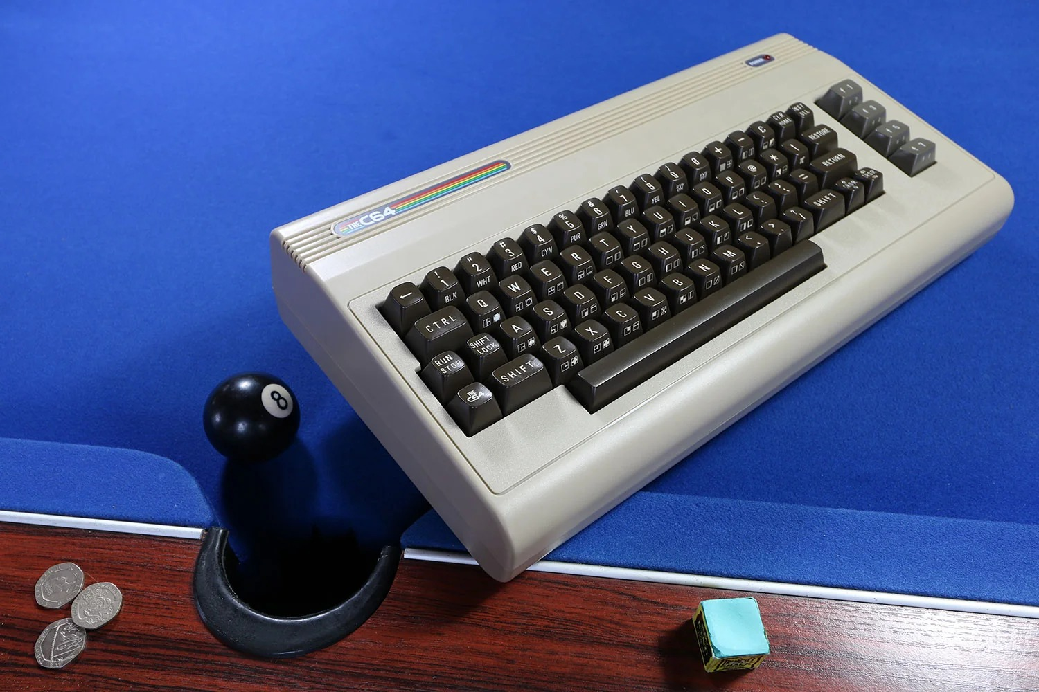 Ein beige Commodore 64 Computer liegt auf einem blauen Billardtisch neben einer Kugel, Kreide und Münzen.