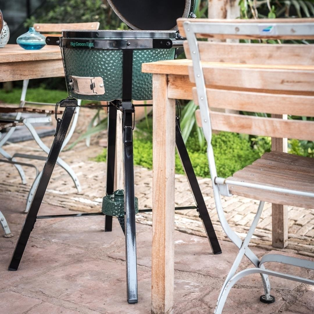 Yeşil renkli Big Green Egg mangal, ahşap mobilyalar arasında dışarıda taş bir verandada duruyor.