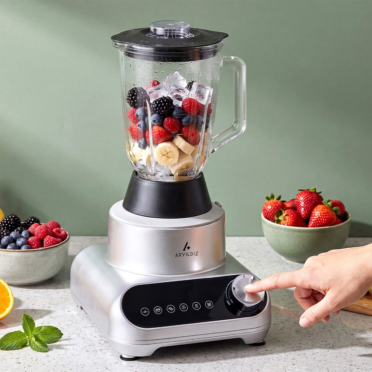 Meyve ve buz dolu gümüş renkli bir blender tezgahta duruyor, bir el düğmeyi çeviriyor.