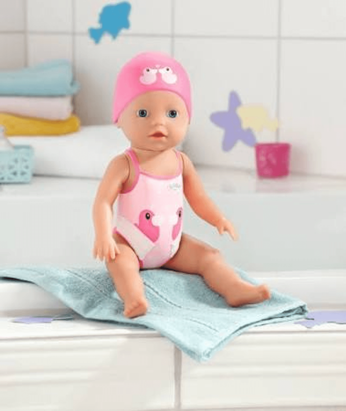 Eine Puppe mit rosa Badeanzug und Badekappe sitzt auf einem blauen Handtuch am Badewannenrand.