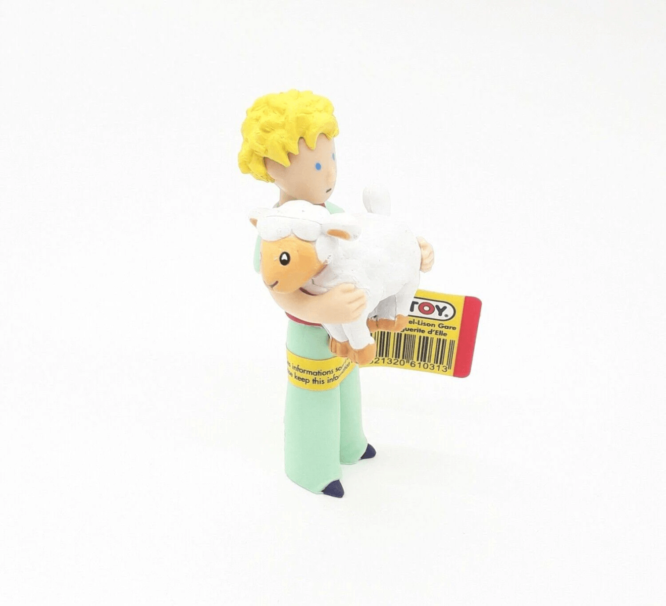 Una pequeña figura del Principito con cabello rubio sostiene una oveja blanca en sus brazos sobre fondo blanco.