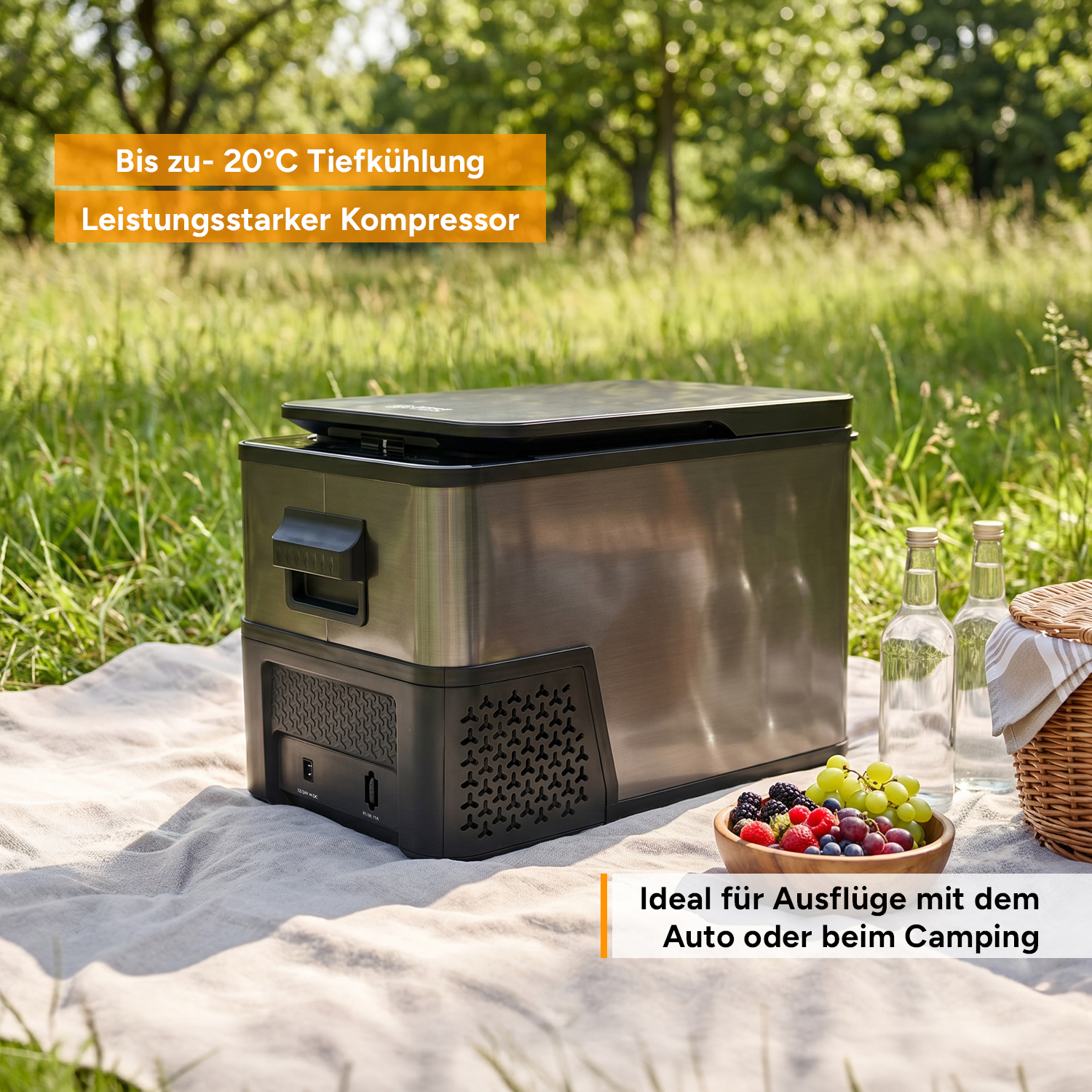Eine tragbare Kompressor-Kühlbox steht auf einer Picknickdecke im Grünen, neben Flaschen, Beeren und einem Korb.