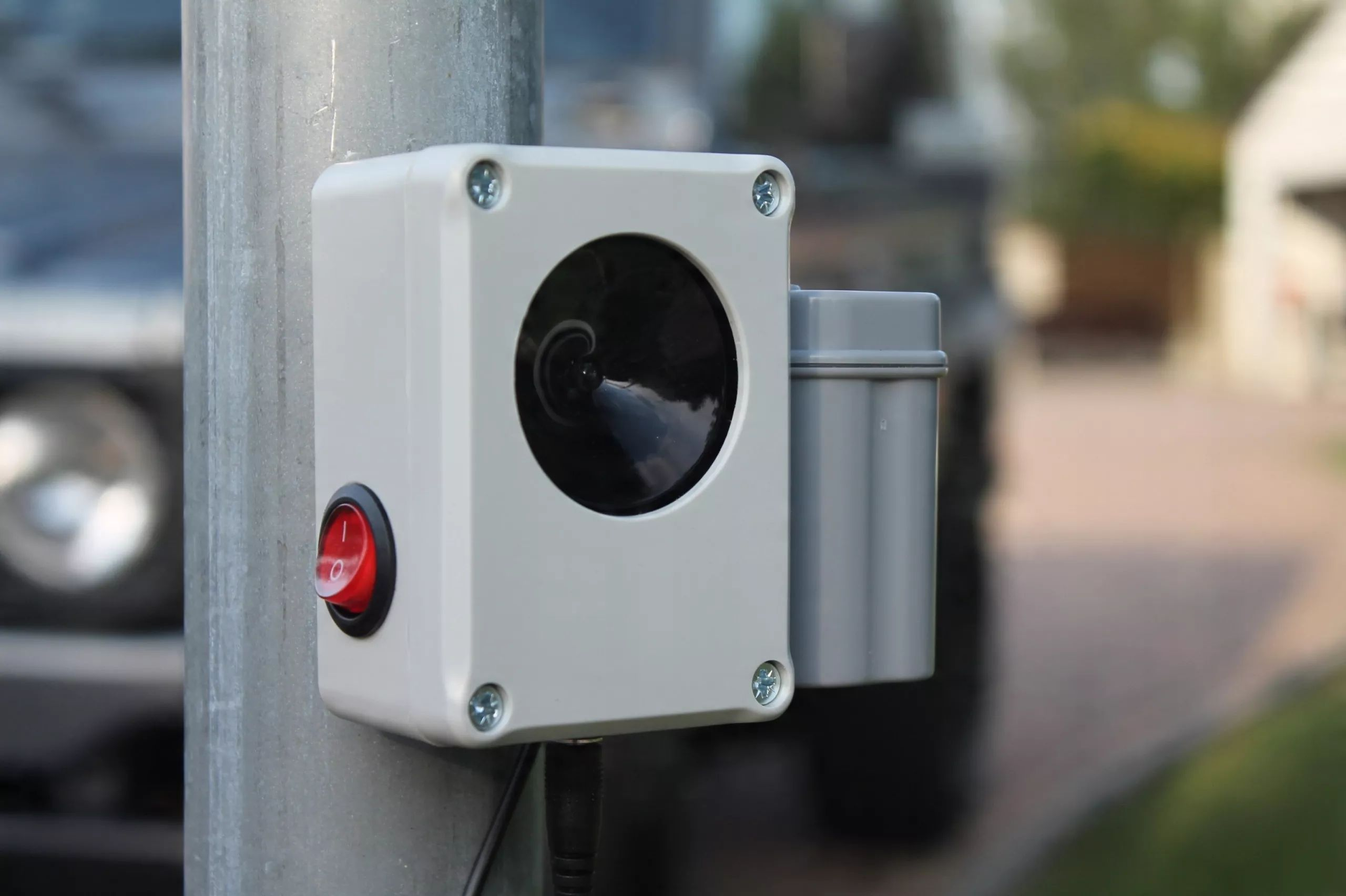 Egy szürke elektronikus doboz fekete hangszóróval és piros kapcsolóval egy fém oszlopra van szerelve.