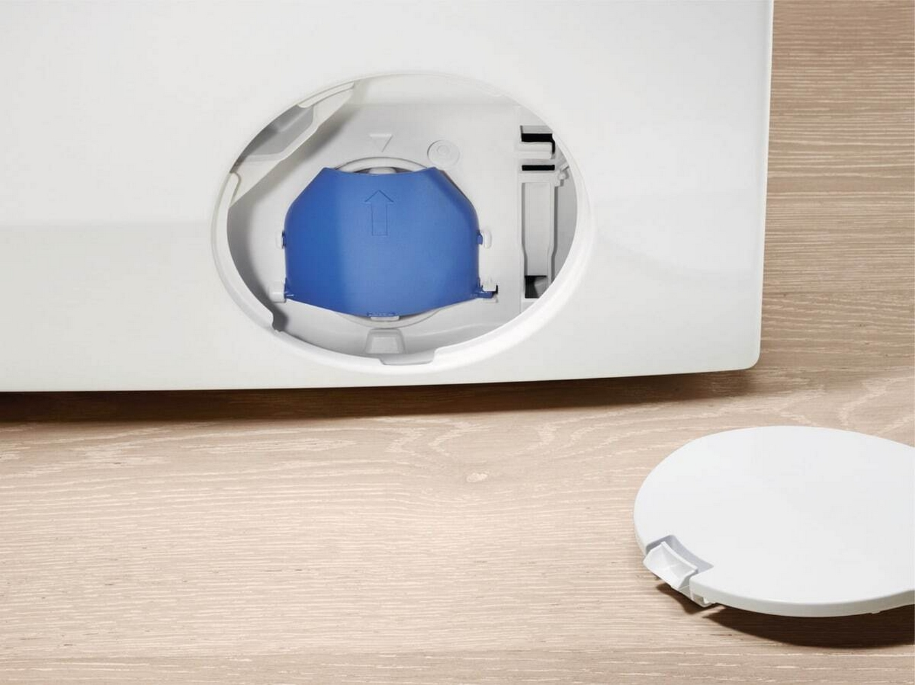 Scomparto filtro lavatrice aperto con tappo blu all'interno e coperchio bianco sul pavimento.
