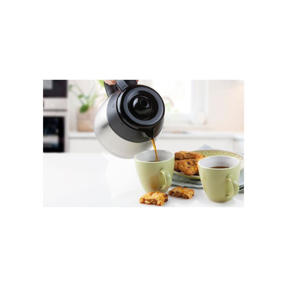 Eine silberne Kaffeekanne gießt Kaffee in eine grüne Tasse. Plätzchen liegen auf dem weißen Tisch in der Küche.