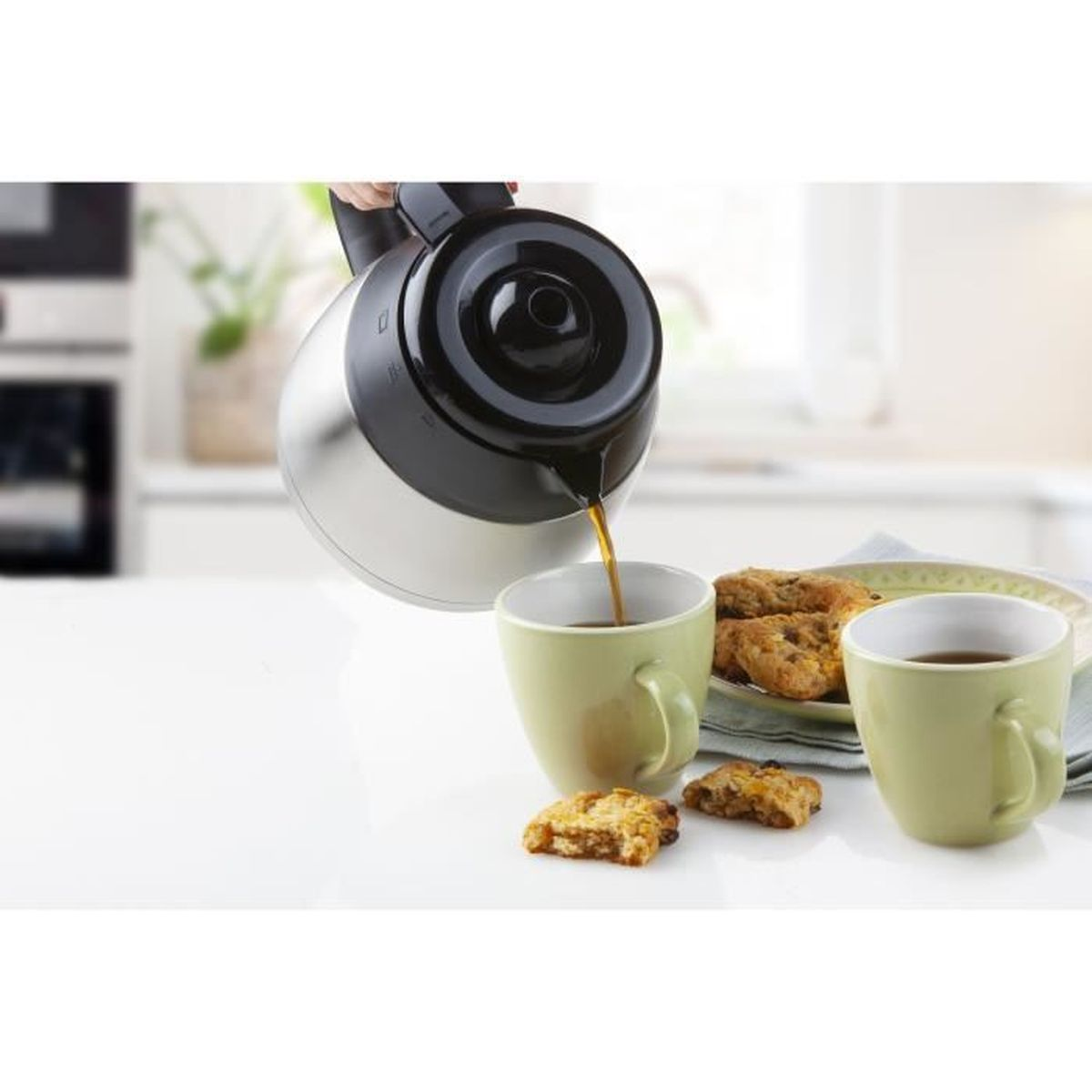 Kaffee wird aus einer silbernen Kanne in eine grüne Tasse gegossen, neben Keksen auf einem Tisch.