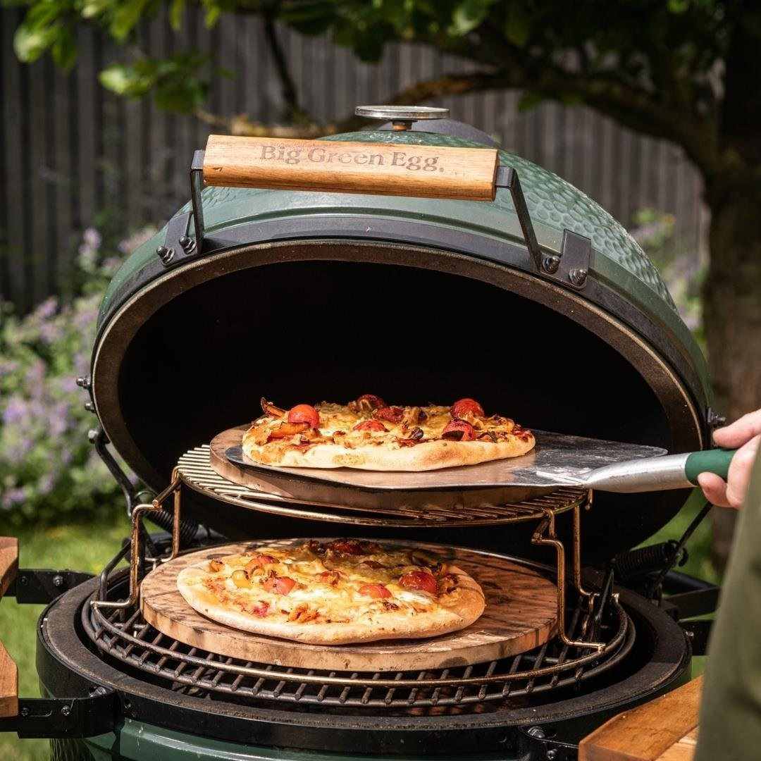 Yuvarlak yeşil Big Green Egg mangalında ahşap tahtalar üzerinde iki pizza pişiriliyor.