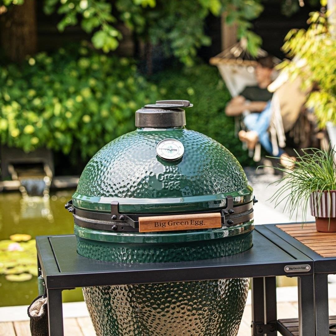 Arka planda havuz ve hamak olan bahçe masasında yeşil seramik barbekü.