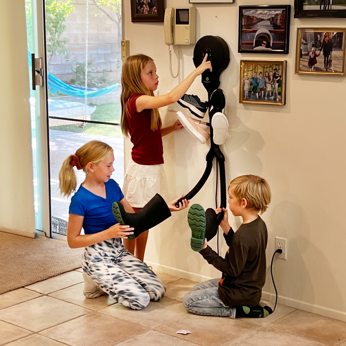 Drei Kinder spielen im Flur mit einem Wandstaubsauger und Schuhen.