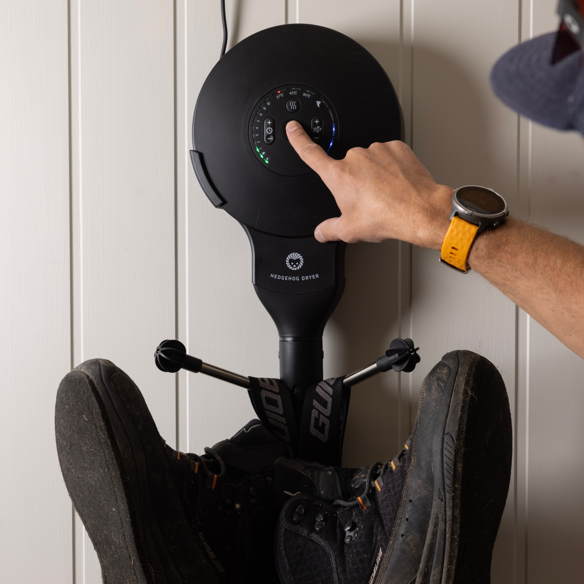 Eine Hand drückt den Ein-Ausschalter eines schwarzen Wand-Stiefeltrockners, der schmutzige Stiefel hält.