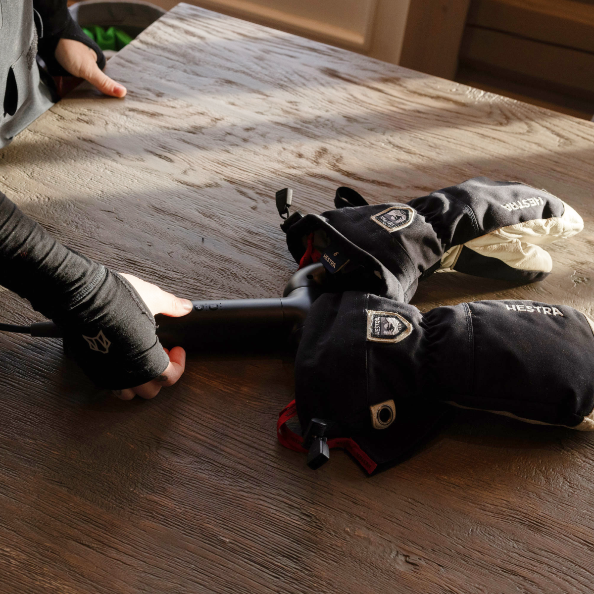 Eine Person benutzt einen Föhn, um schwarze Hestra Skihandschuhe auf einem Holztisch zu trocknen.
