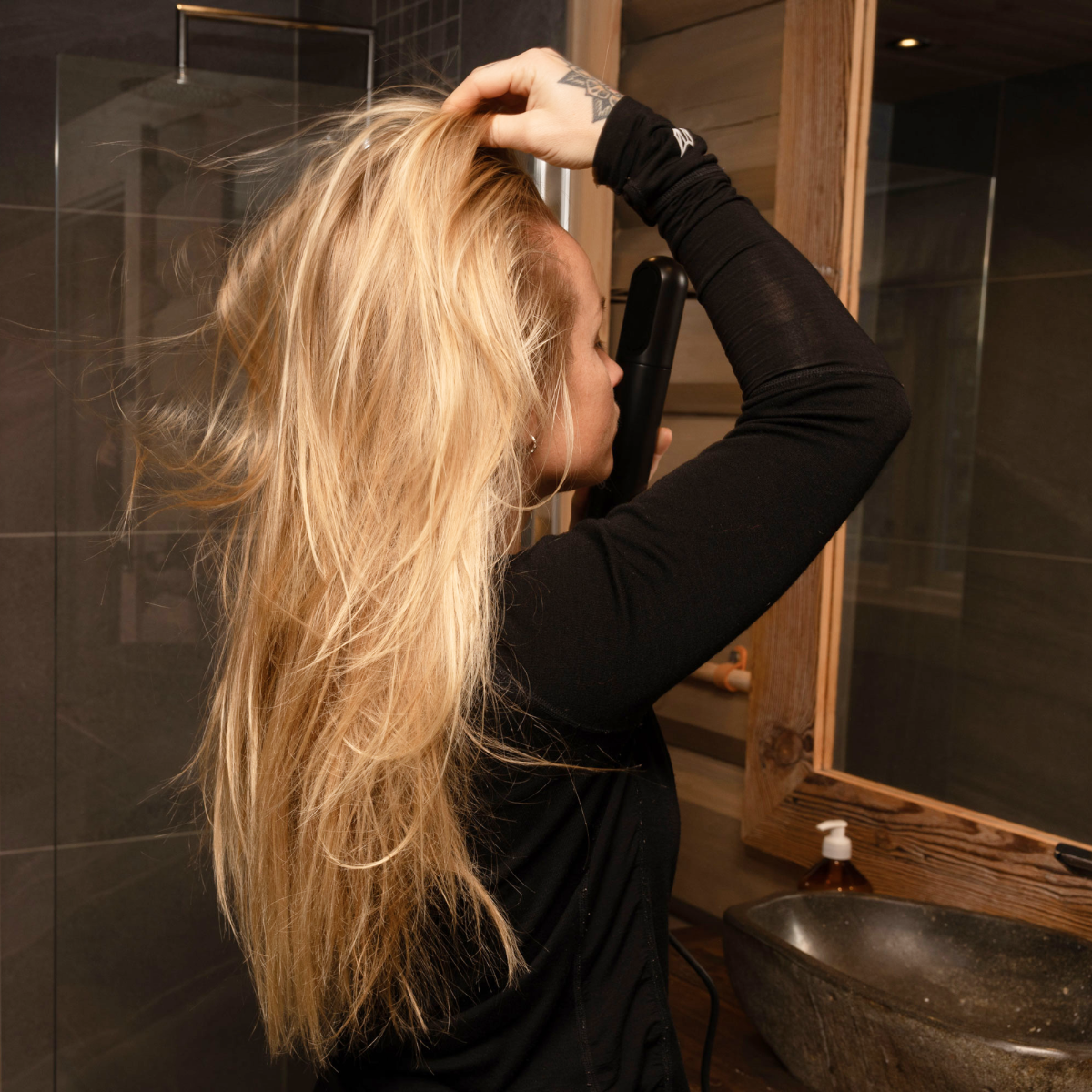 Eine blonde Frau im schwarzen Oberteil glättet sich die Haare im Badezimmer vor einem Spiegel.