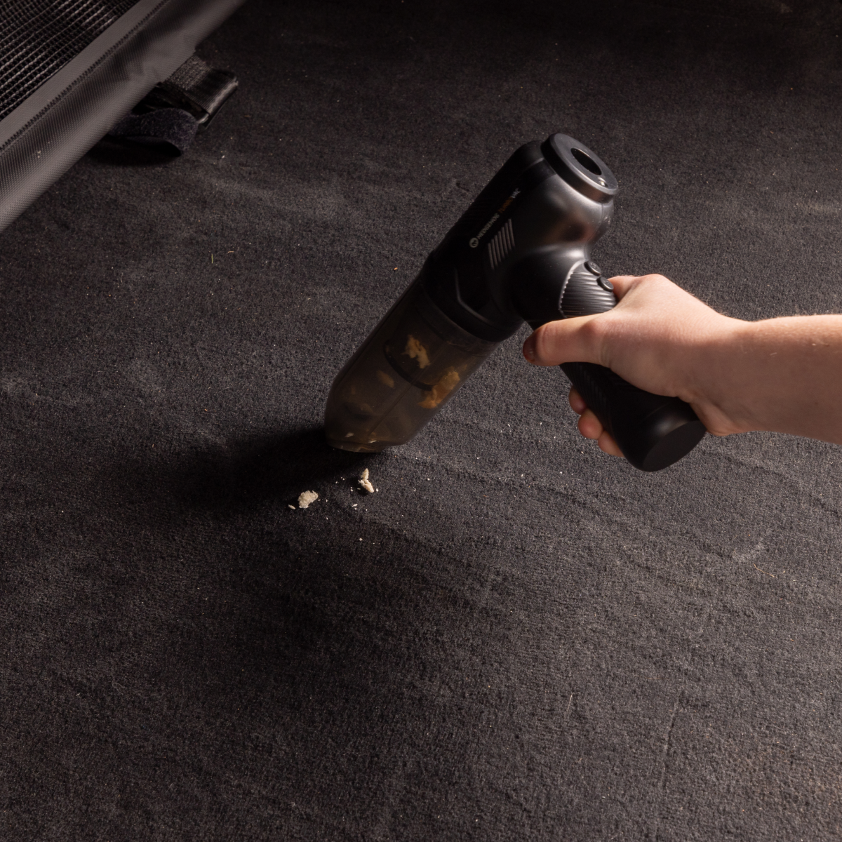 Eine Hand saugt Krümel mit einem kleinen schwarzen Handstaubsauger von dunklem Autoteppich auf.