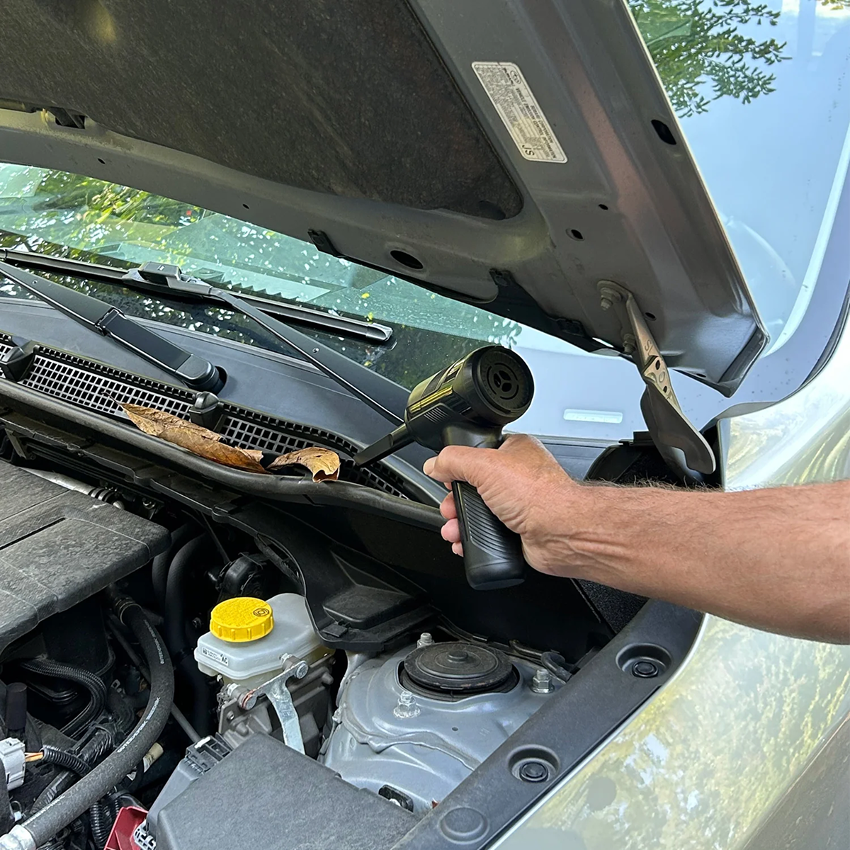 Person hält ein kleines schwarzes Werkzeug in einem offenen Auto-Motorraum mit Laub.