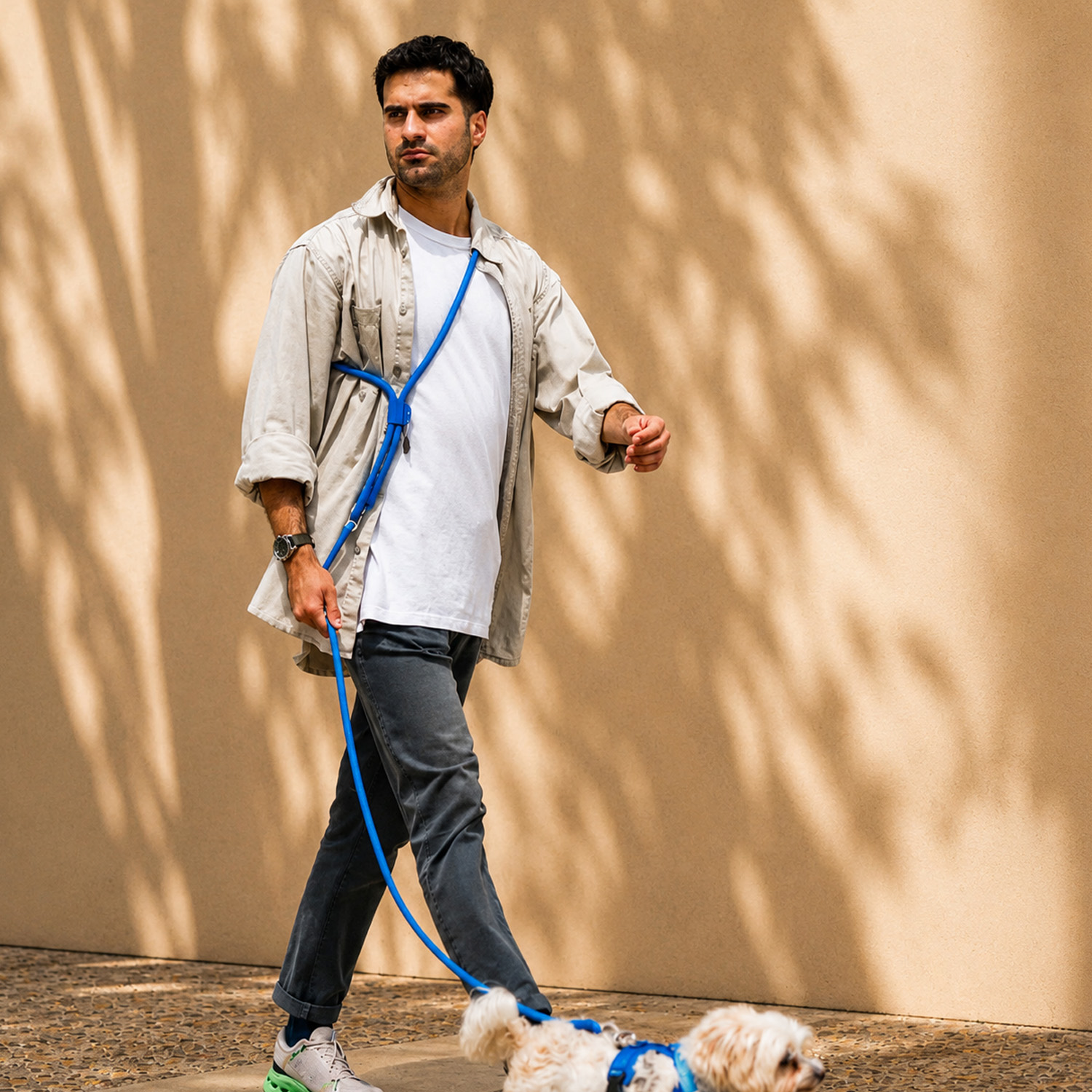 Un hombre con camisa blanca pasea un perro blanco pequeño con correa azul frente a una pared.