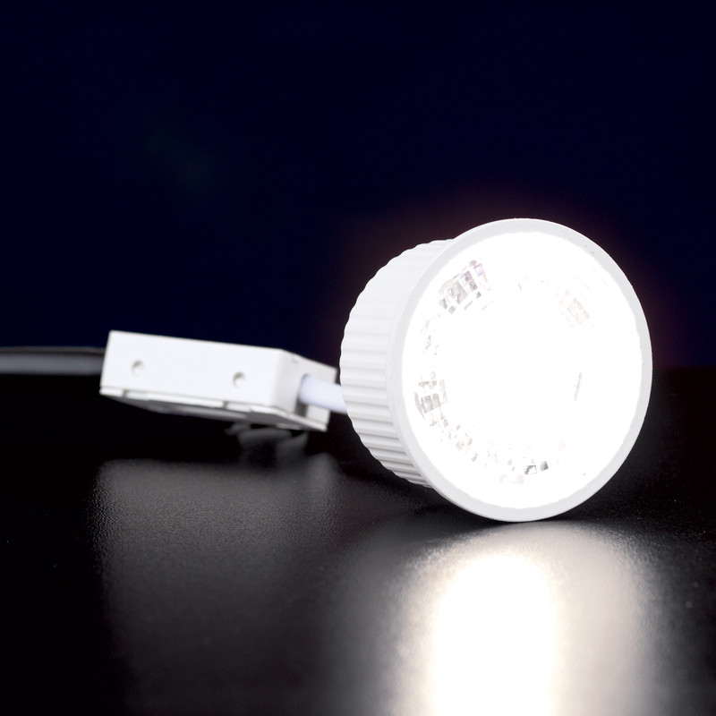 Ein heller weißer LED-Strahler liegt auf einer dunklen Oberfläche und ist mit einer kleinen weißen Anschlussbox verbunden.