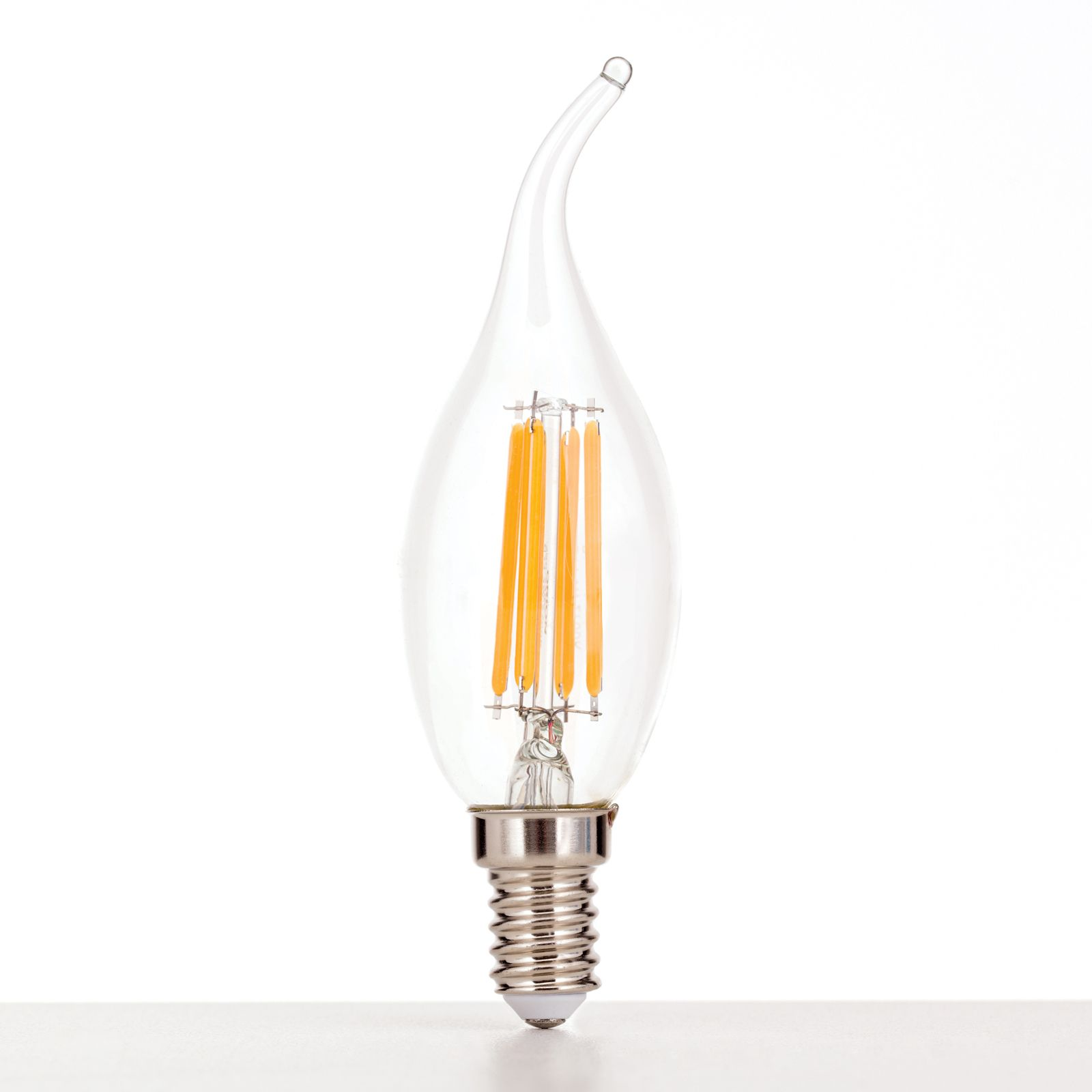 Eine geschwungene LED-Kerzenlampe mit sichtbaren Fäden steht auf einer weißen Oberfläche vor hellem Hintergrund.