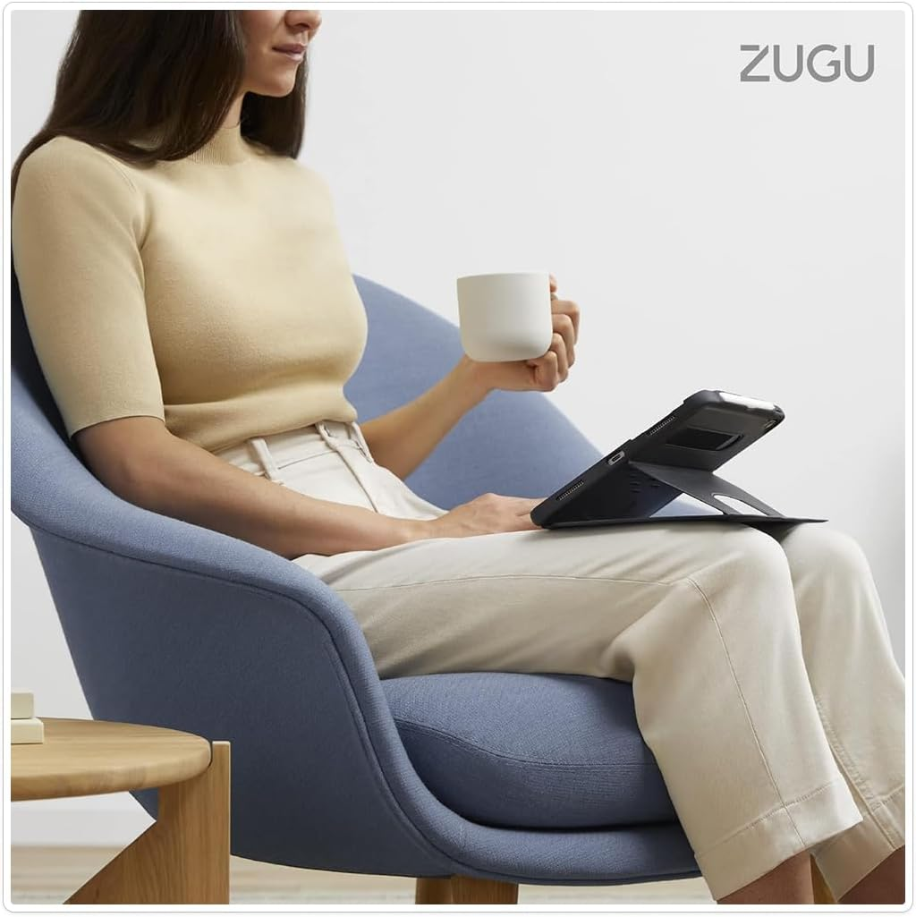 Eine Frau in beige Kleidung sitzt auf einem blauen Sessel, hält eine Tasse und nutzt ein Tablet auf ihren Beinen.