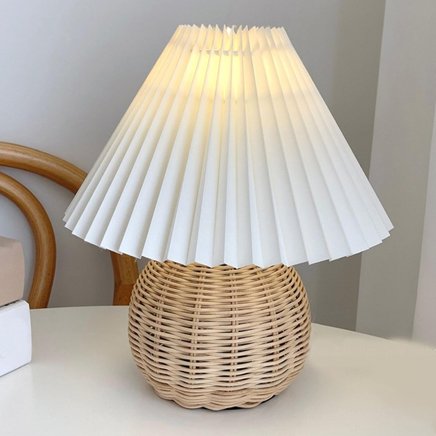 Eine Tischlampe mit rundem Korbsockel und weißem Plissérschirm auf weißem Tisch.