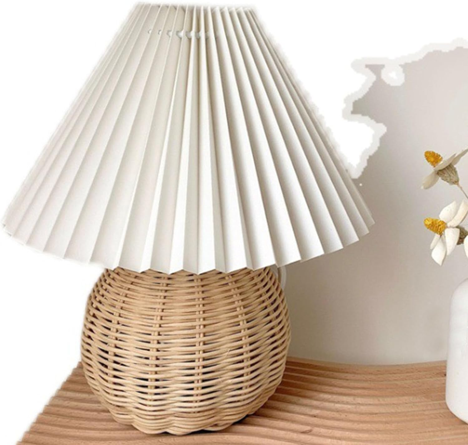 Eine Tischlampe mit rundem Rattansockel und weißem, plissiertem Schirm auf einer Holzfläche.