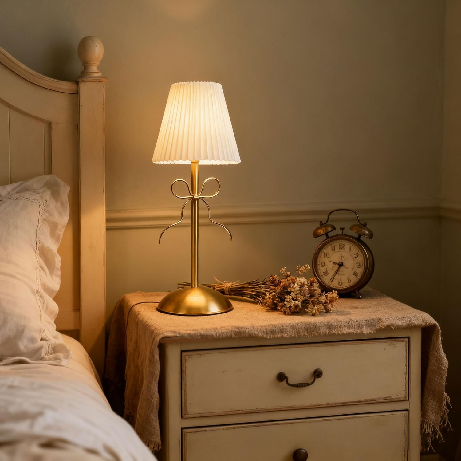 Eine beleuchtete Plissé-Lampe, Trockenblumen und eine alte Uhr stehen auf einem weißen Nachttisch.