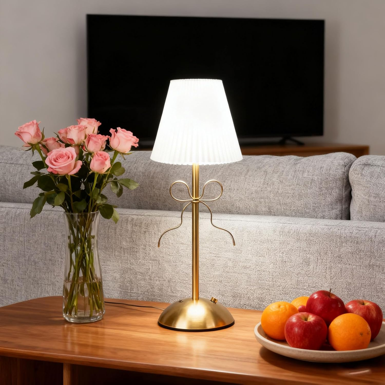 Eine beleuchtete Goldlampe, eine Vase mit rosa Rosen und eine Obstschale auf einem Holztisch, dahinter ein graues Sofa und ein Fernseher.