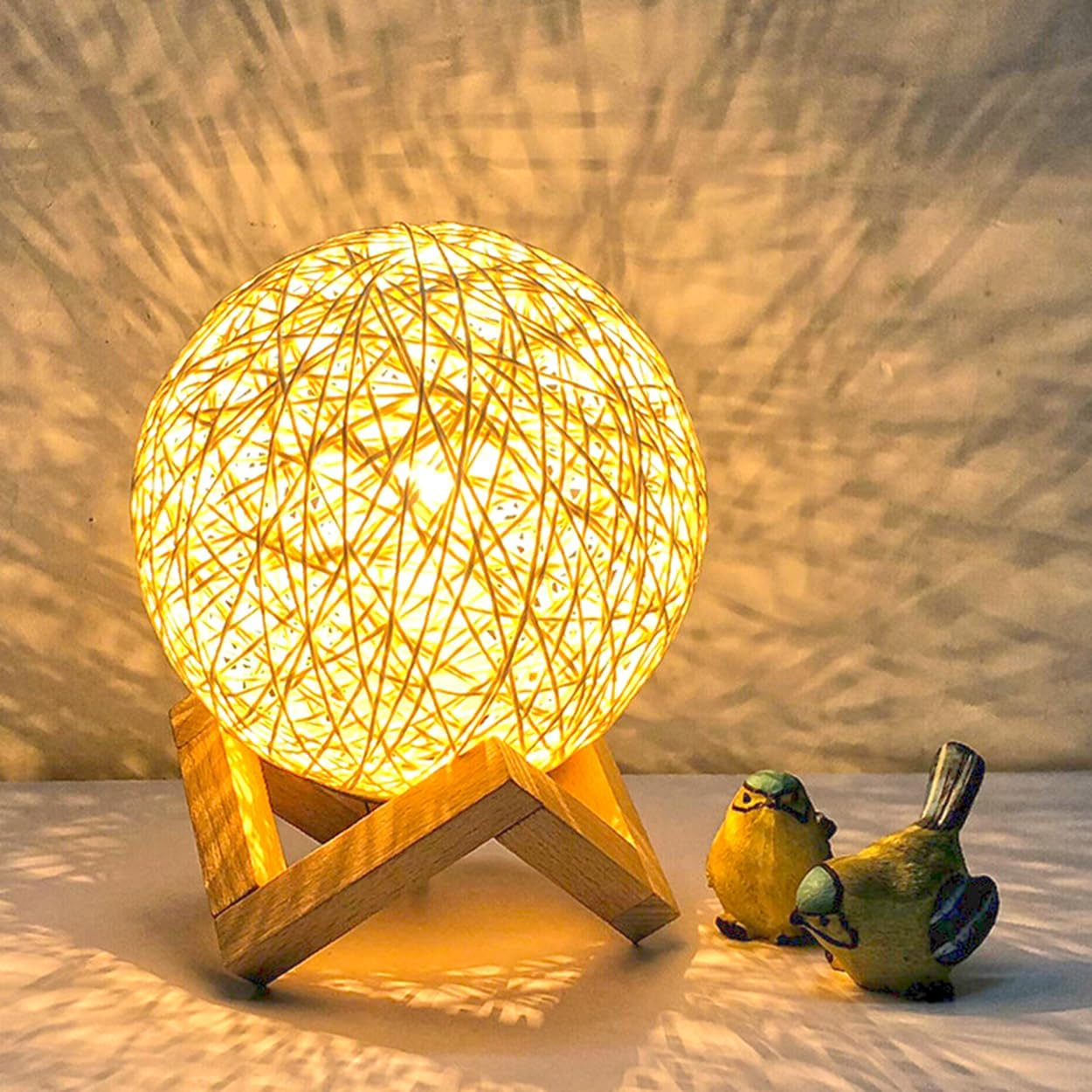 Eine leuchtende Kugel aus Rattan auf Holzständer mit zwei kleinen Vogelstatus daneben.